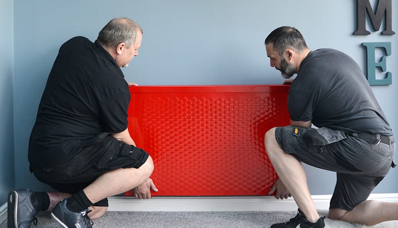Two Sunflow installers installing a red Sunflow classic radiator on a customer's wall in a light blue or grey room.