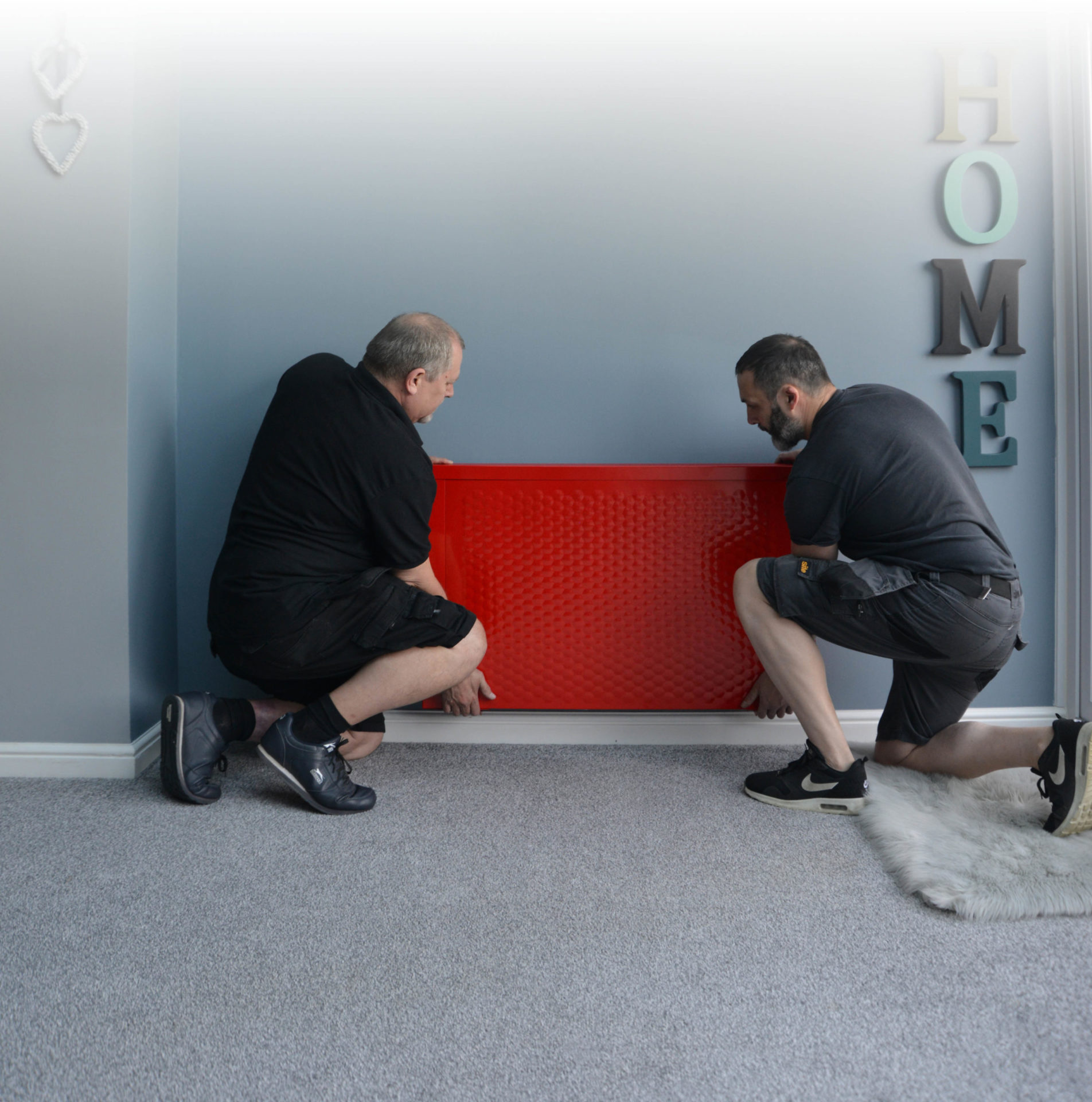 Two Sunflow installers installing a red Sunflow classic radiator on a light blue or grey wall of a customer's home.