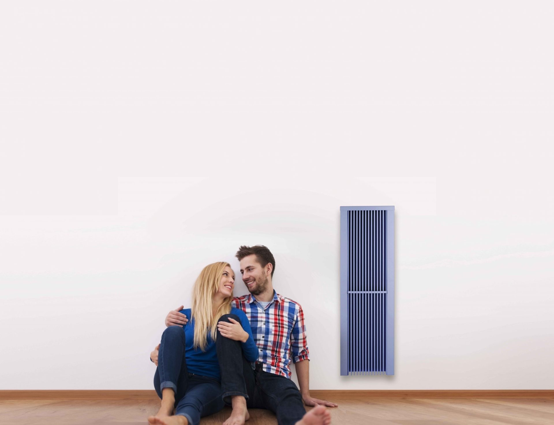Young couple sitting on floor next to a blue Sunflow classic radiator, against a white wall with wood flooring.